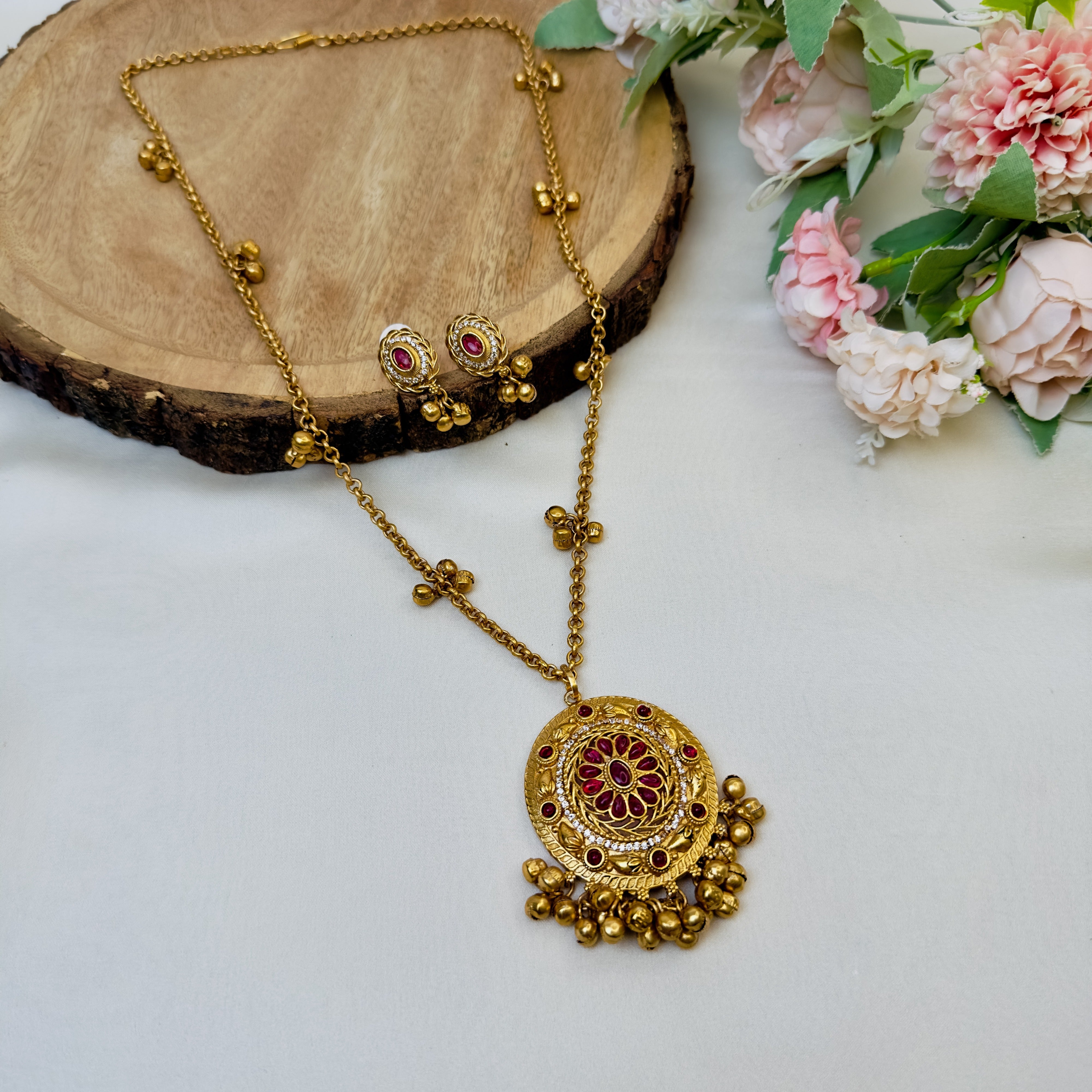 A gold-colored Ghungroo Mala Pendant Set with intricate designs and ornamental details, displayed on a wooden surface with flowers in the background.